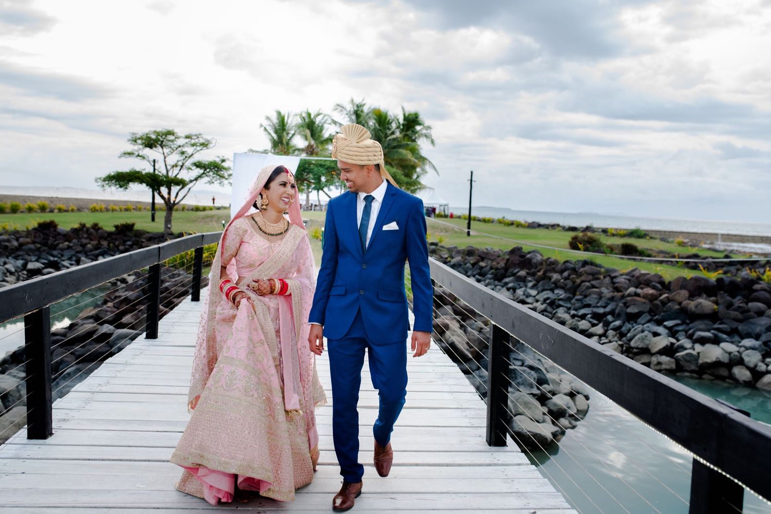 Couple in wedding attire walks along an oceanside boardwalk with palm trees and rocky coast in background.