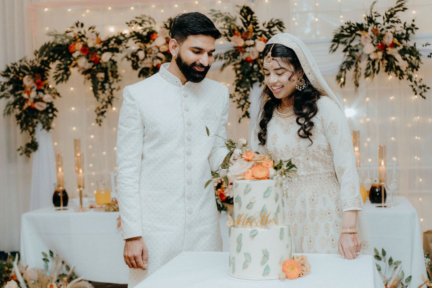 A wedding couple in matching white traditional attire stands together against a floral backdrop with candles.