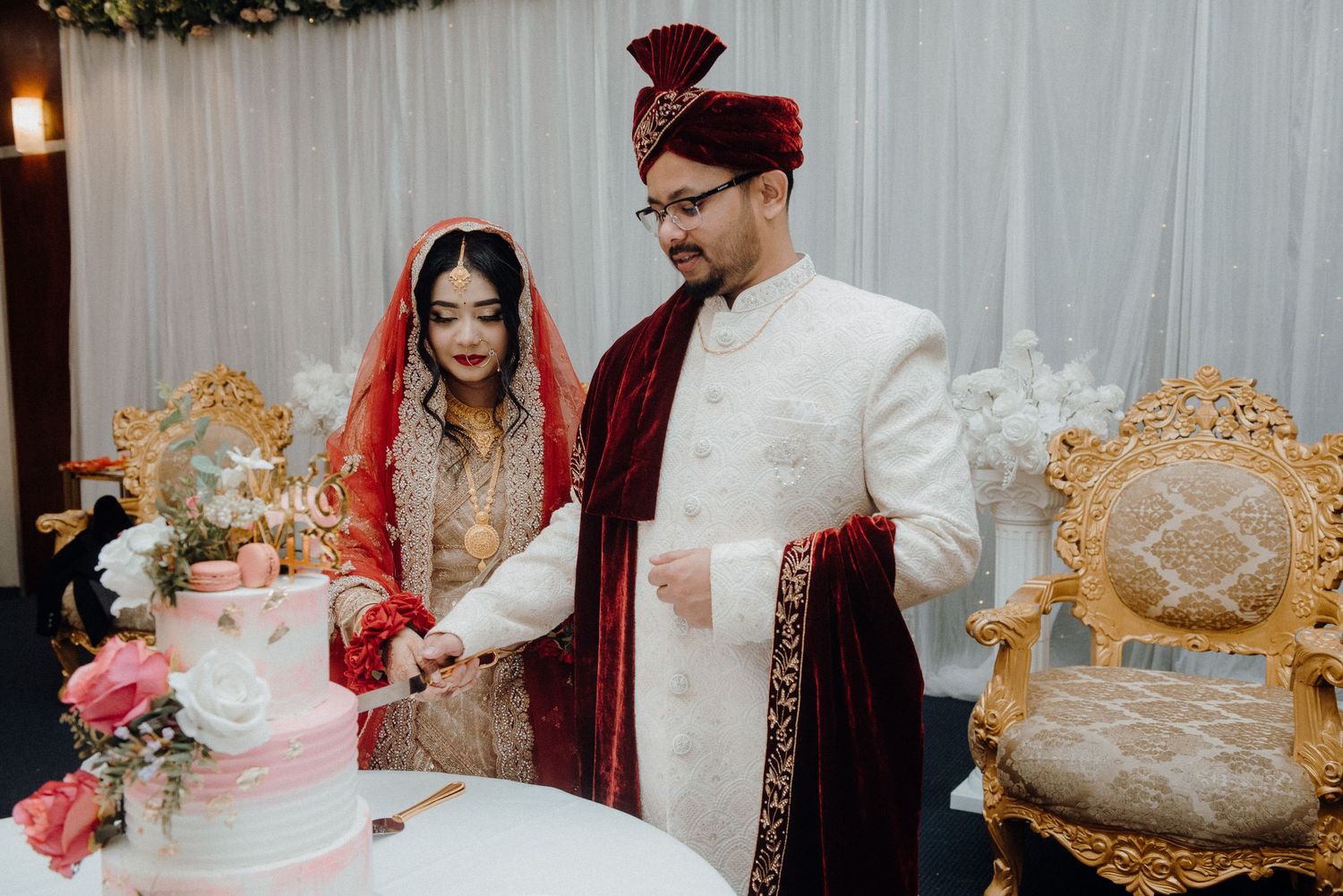 Couple cutting their pink and white tiered wedding cake beside ornate gold chairs.