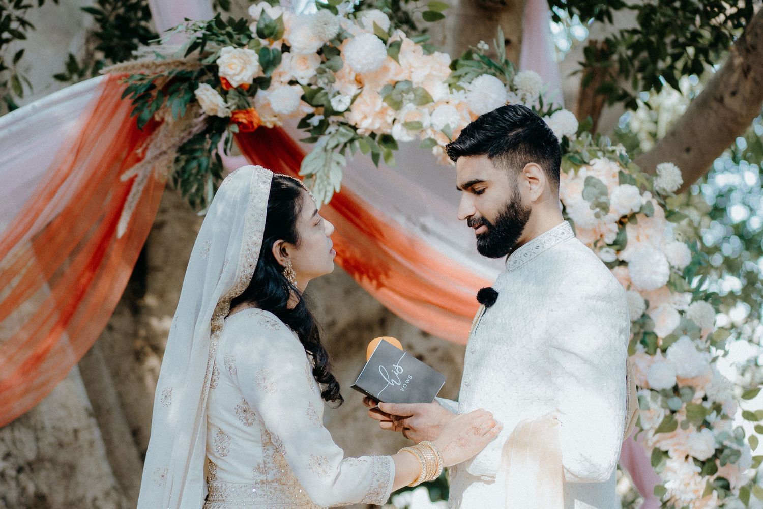 A couple exchanges vows under a floral arch with orange draping and white flowers.