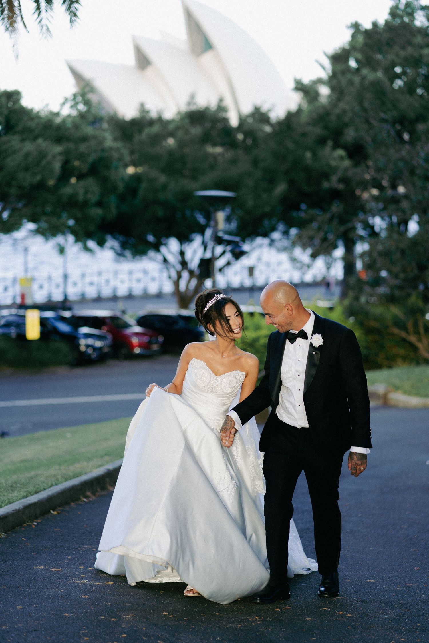Wedding couple walking near Sydney Opera House, bride in white gown and groom in black tuxedo.