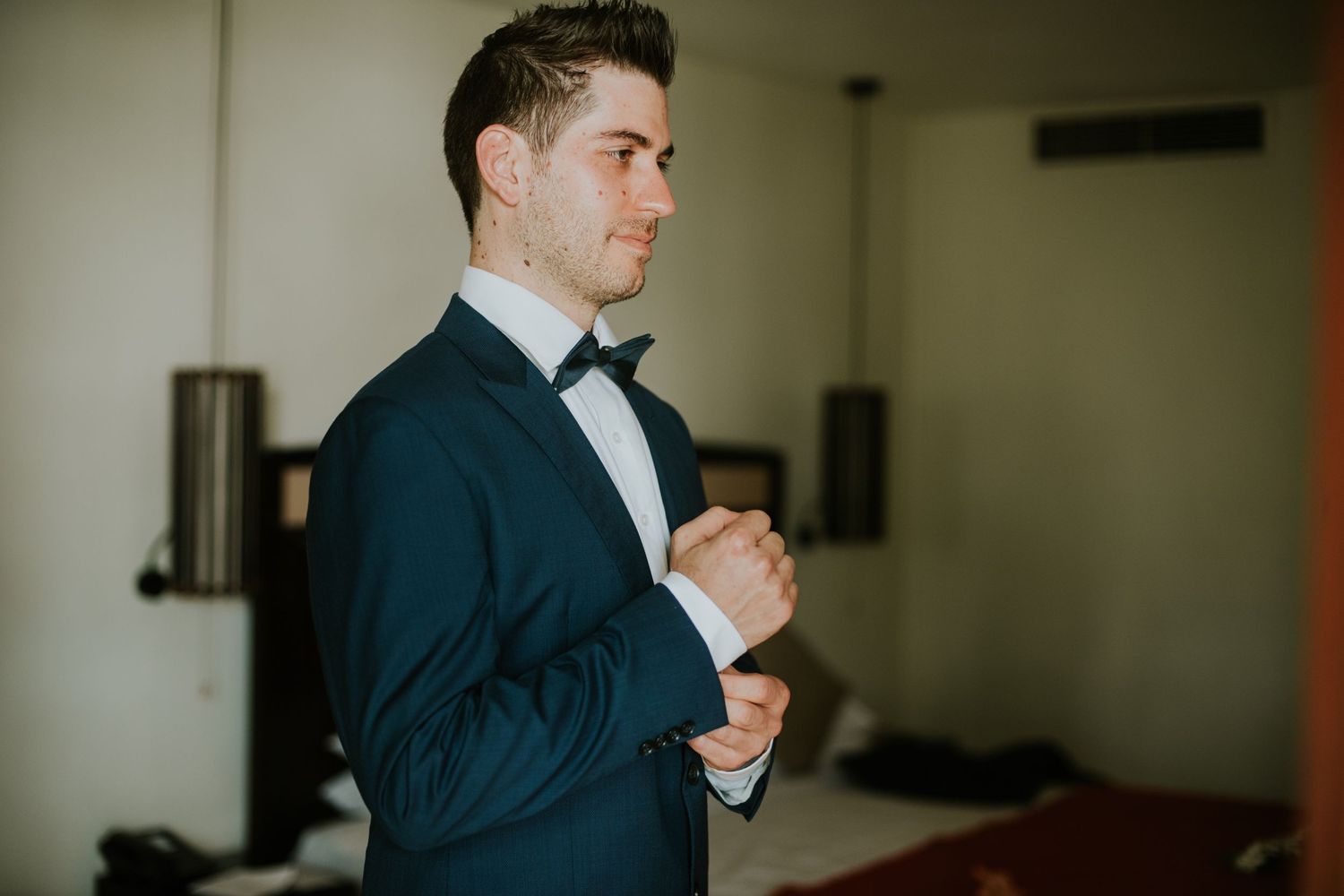 Groom adjusting bowtie while wearing navy tuxedo in hotel room.