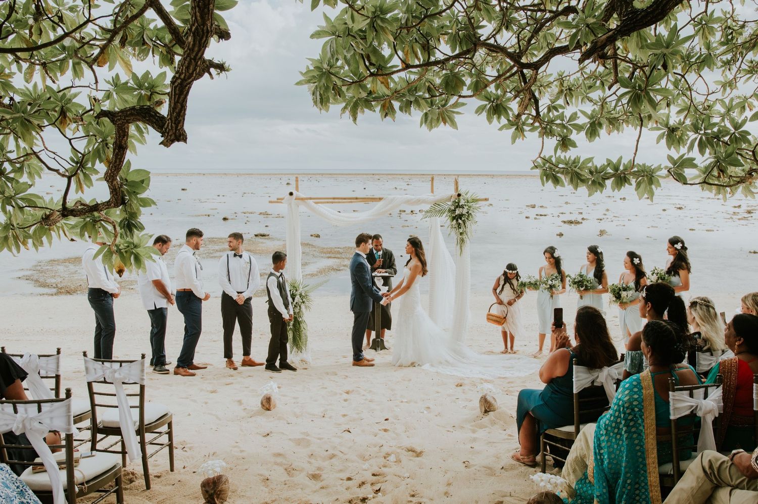 Guests gather on a beach for an outdoor wedding ceremony with palm trees and ocean views.