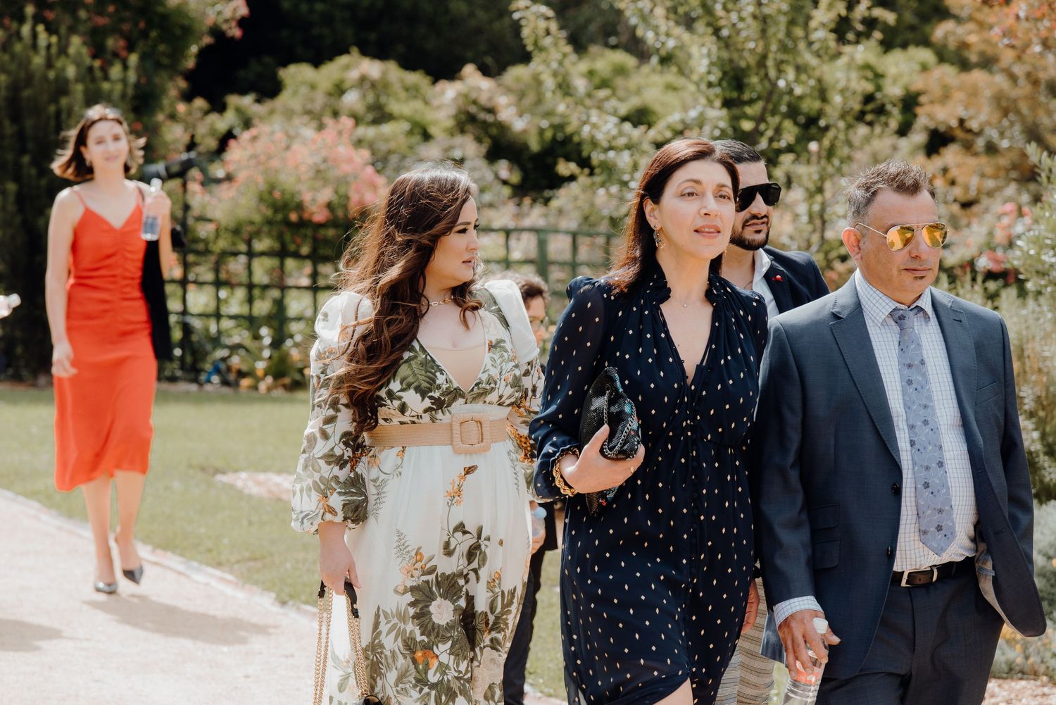 Wedding guests walk together on a sunny garden path dressed in formal attire.