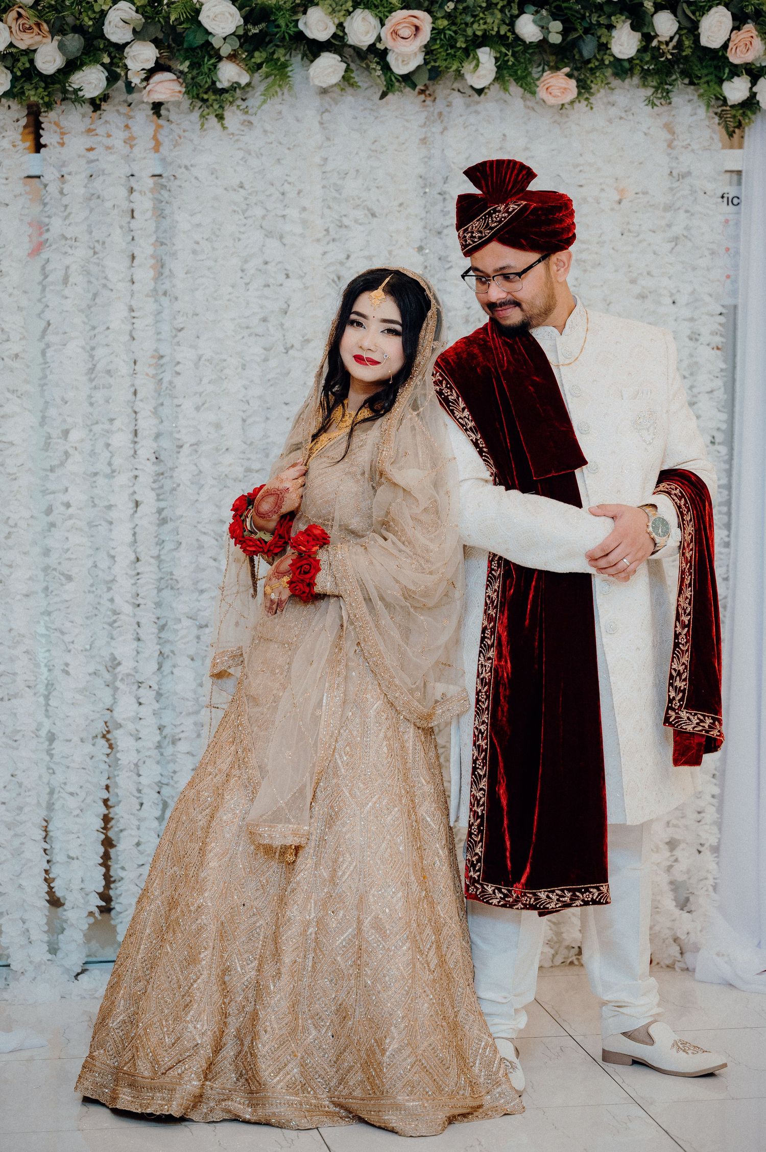 Couple in traditional South Asian wedding attire posing against floral backdrop.