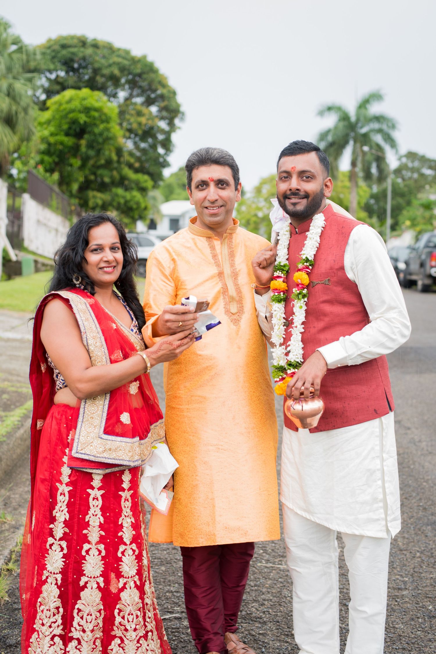 Three people in traditional Indian attire posing together outdoors with palm trees in background.