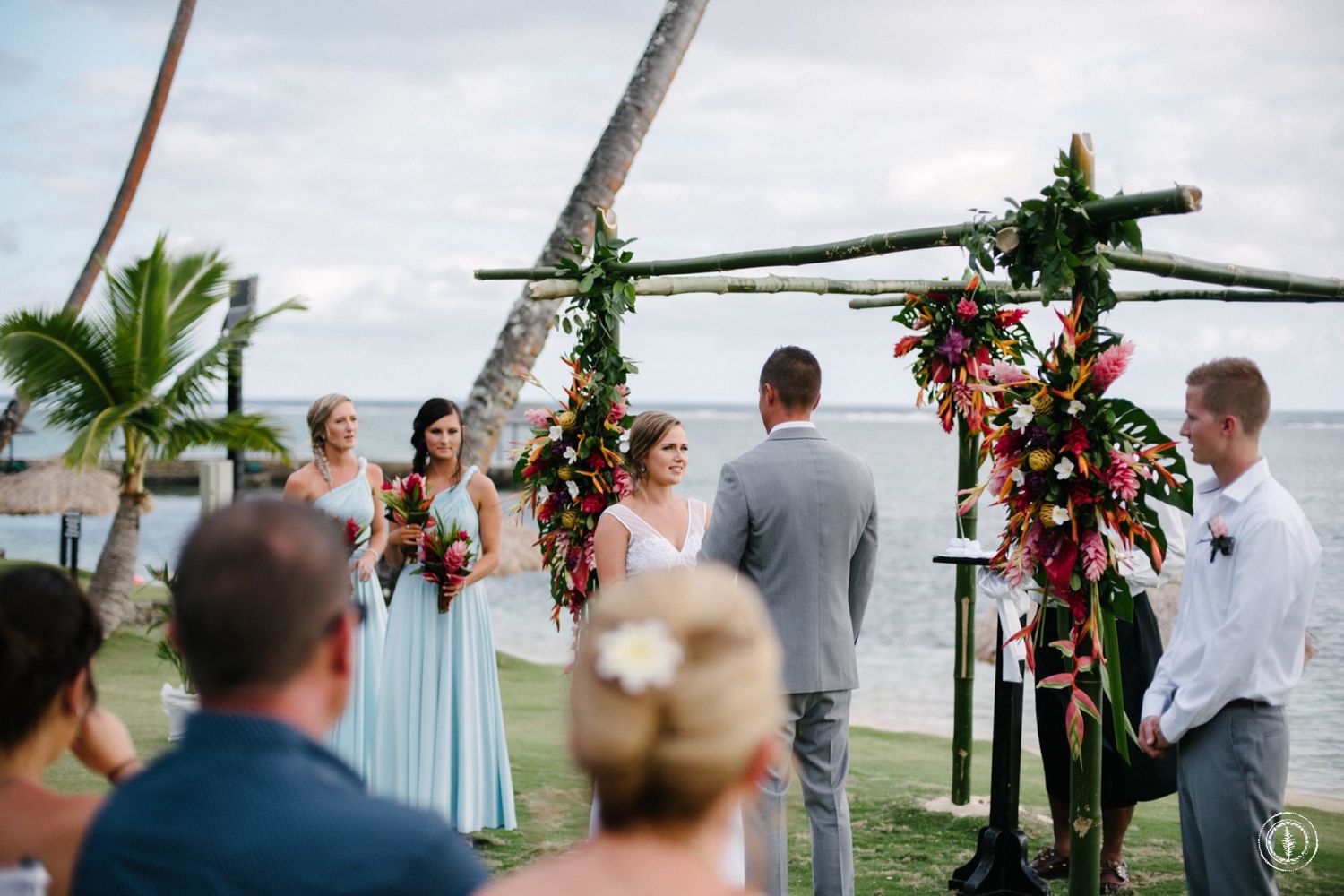Outdoor beach wedding ceremony under a wooden arch decorated with tropical flowers and greenery.