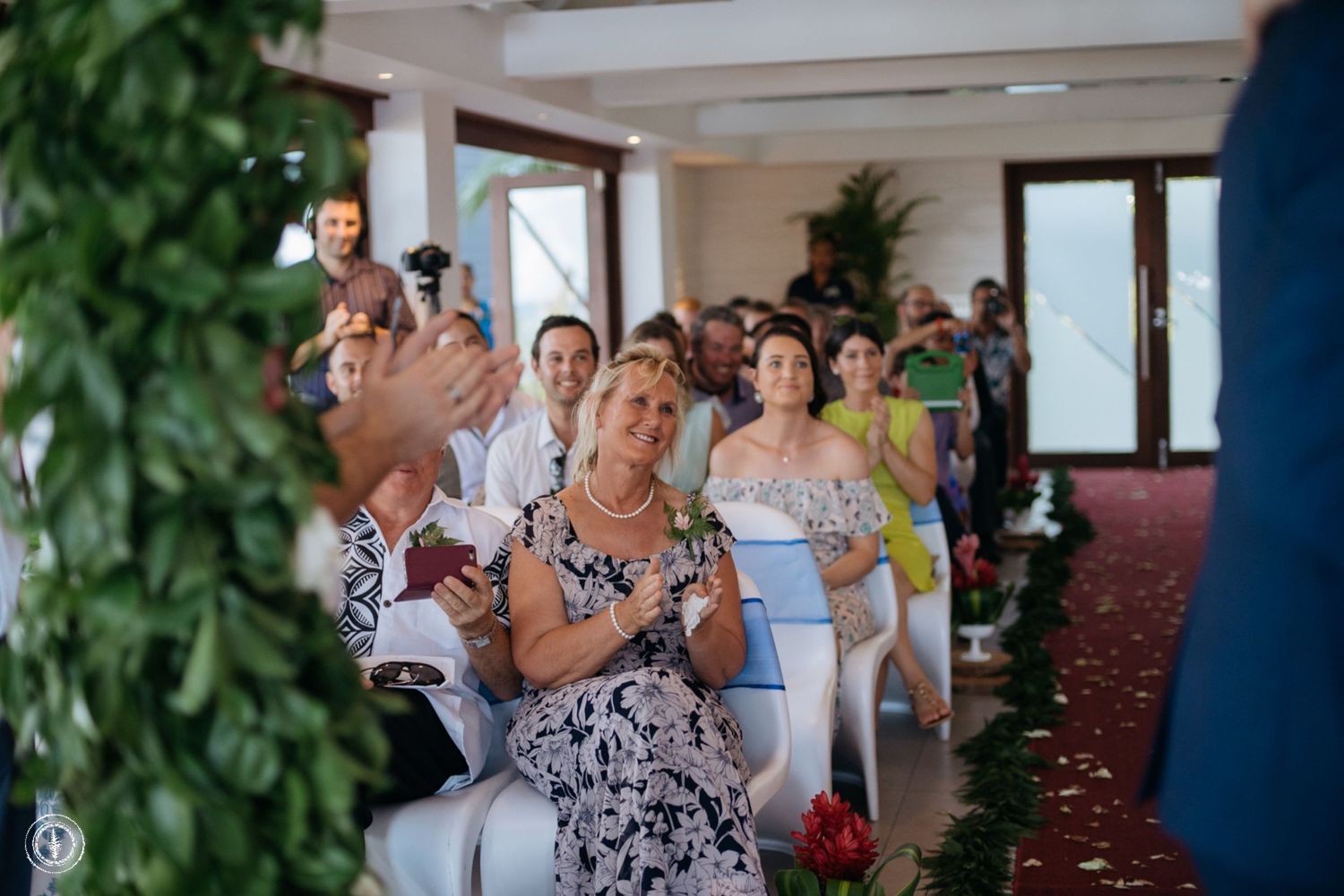 Wedding guests seated in rows at an indoor ceremony applaud with joyful expressions.