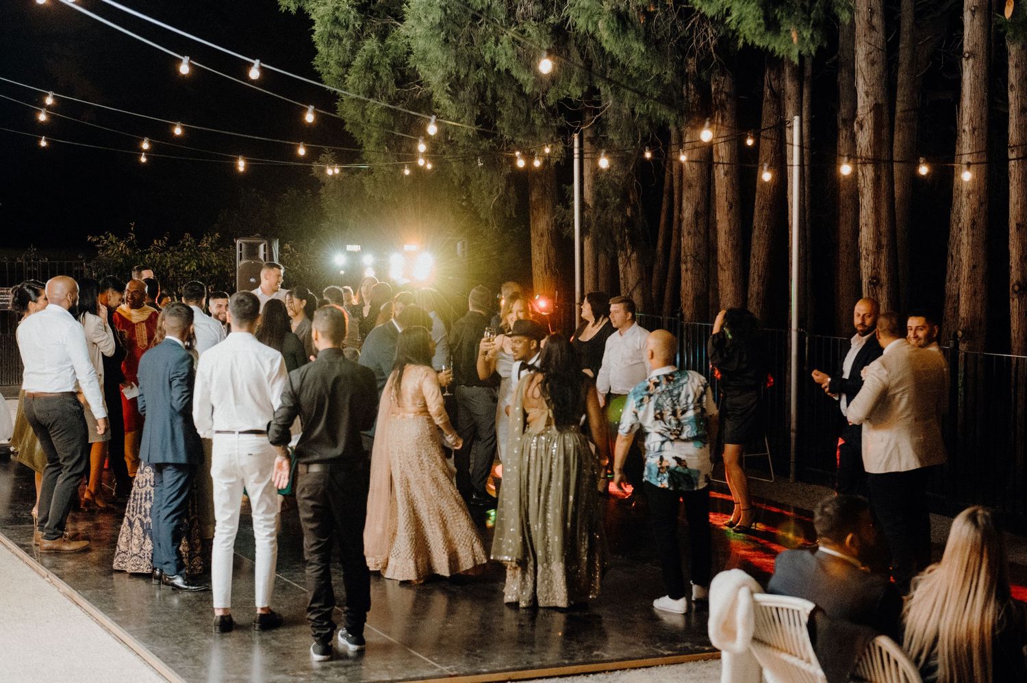 Wedding guests mingling outdoors under string lights at dusk among tall trees.