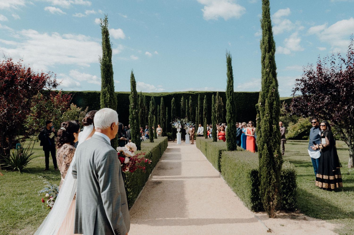 Wedding guests gathering along a cypress-lined garden pathway on a sunny day.