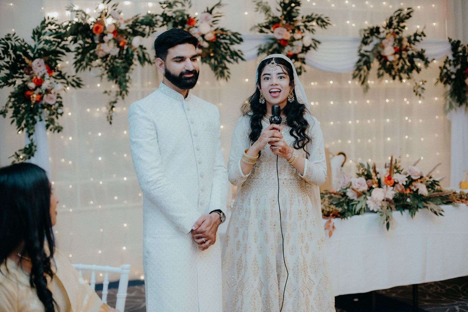 Wedding couple in traditional white attire speaking in front of floral backdrop with twinkle lights.