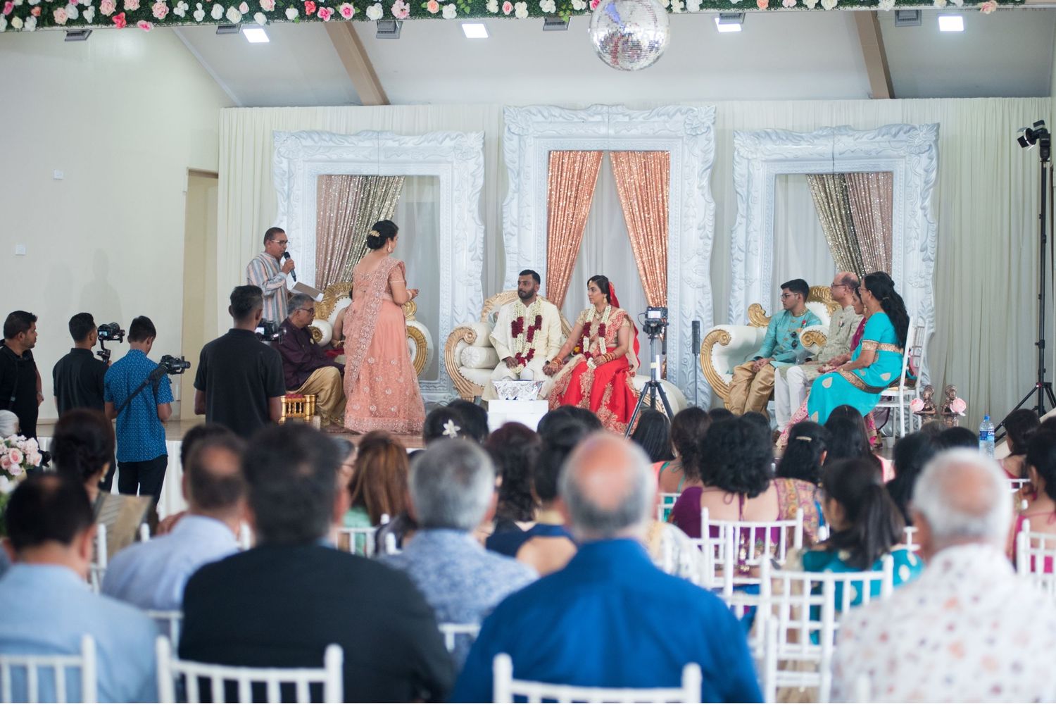 Wide view of a wedding ceremony with guests seated in white chairs facing the decorated stage.