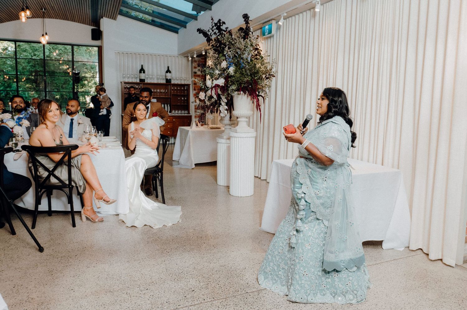 Wedding celebration with speaker in blue dress addressing seated guests in bright venue.