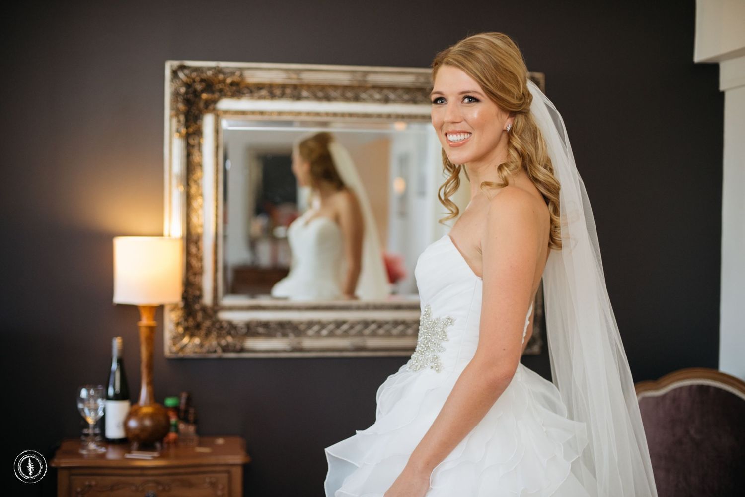 Bride in white wedding gown poses in front of mirror in elegant hotel room setting.