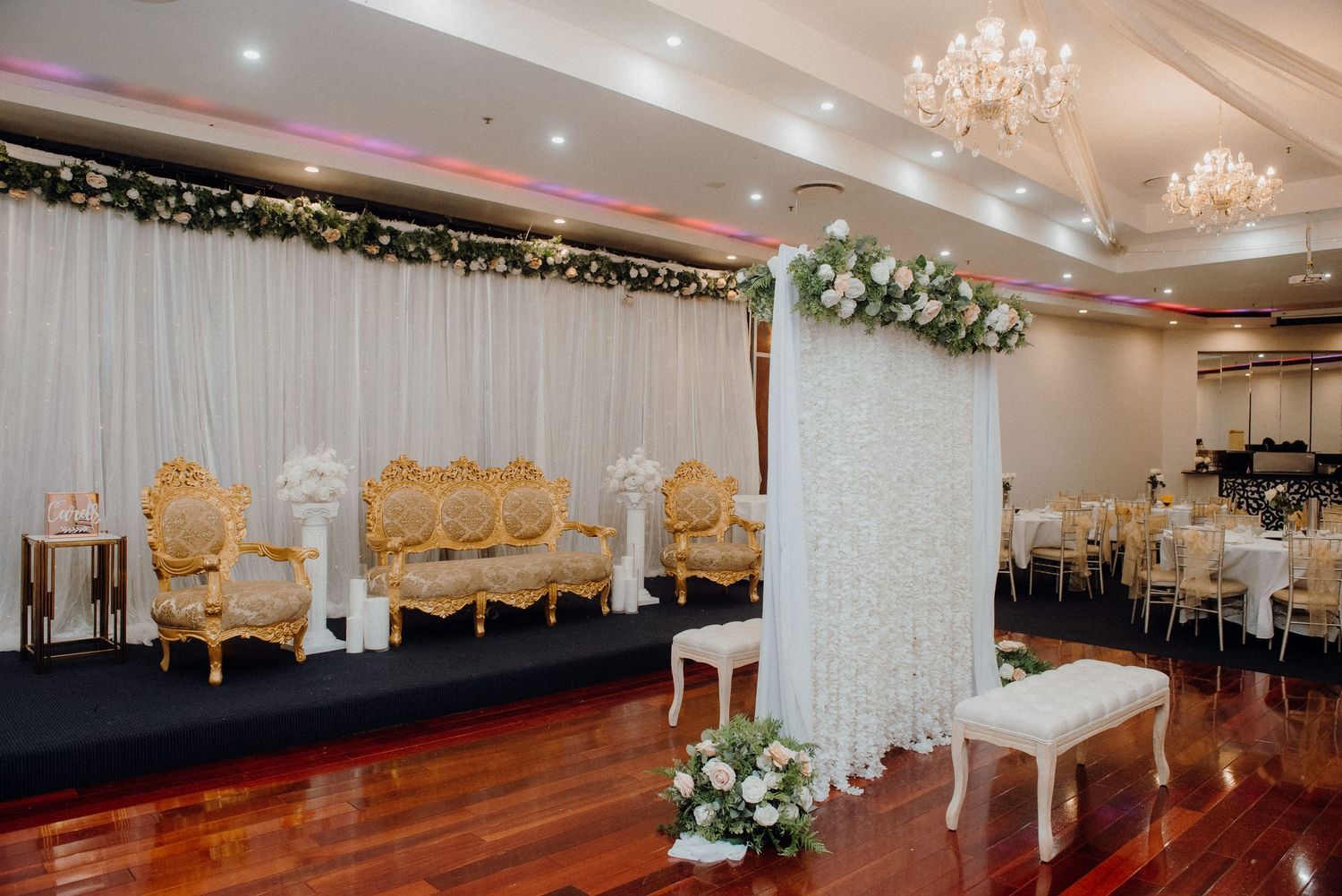Ornate gold seating arrangement with white floral backdrop and decorative pillars in a banquet hall.