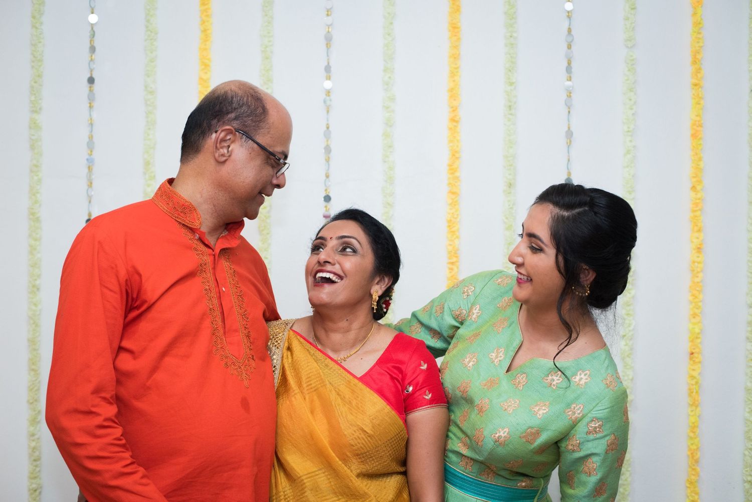 Three people sharing a candid laugh together while wearing traditional Indian attire in orange, yellow and green.