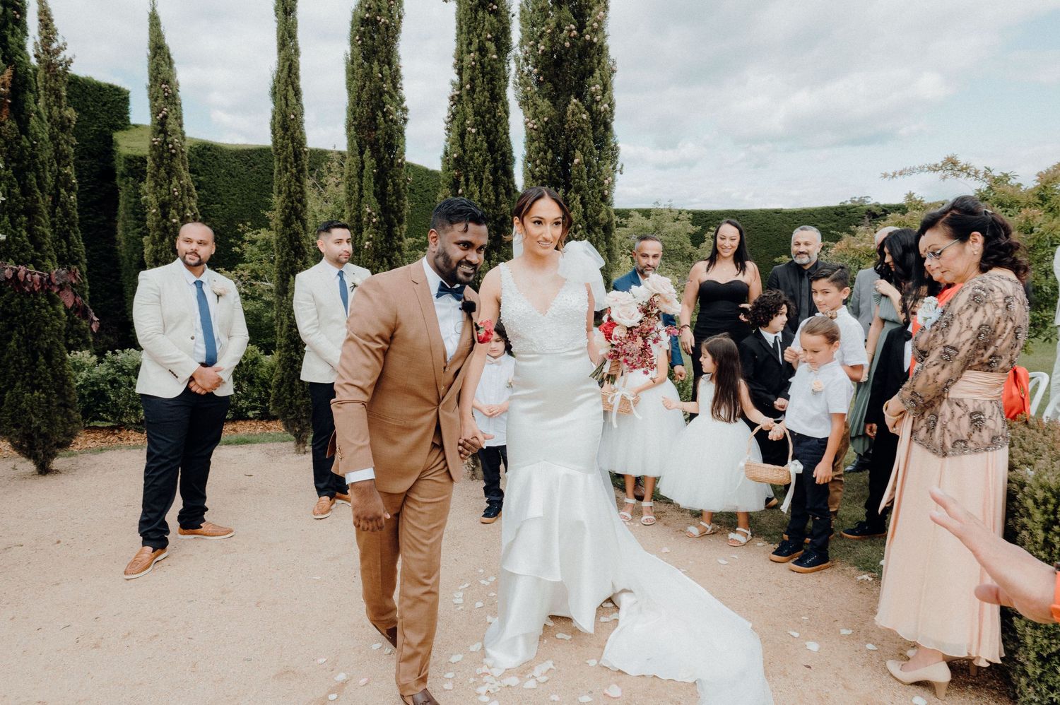 Wedding procession walking along garden path with cypress trees and guests.