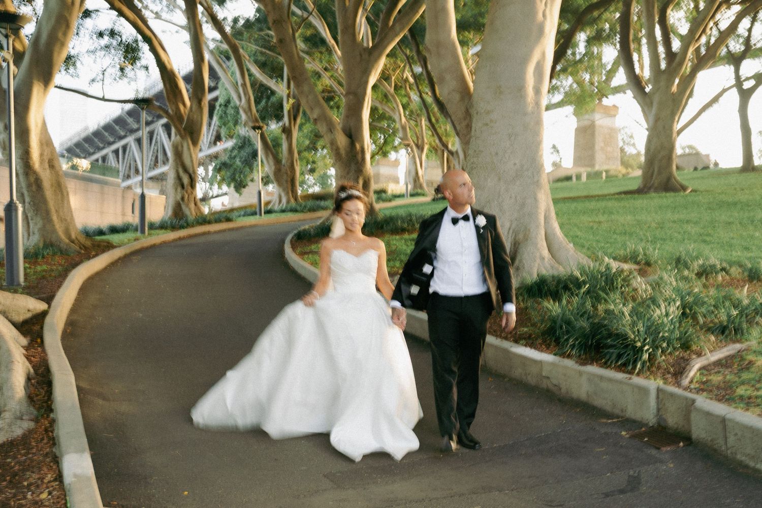 Bride in voluminous white gown and groom in tuxedo walk along tree-lined park path.