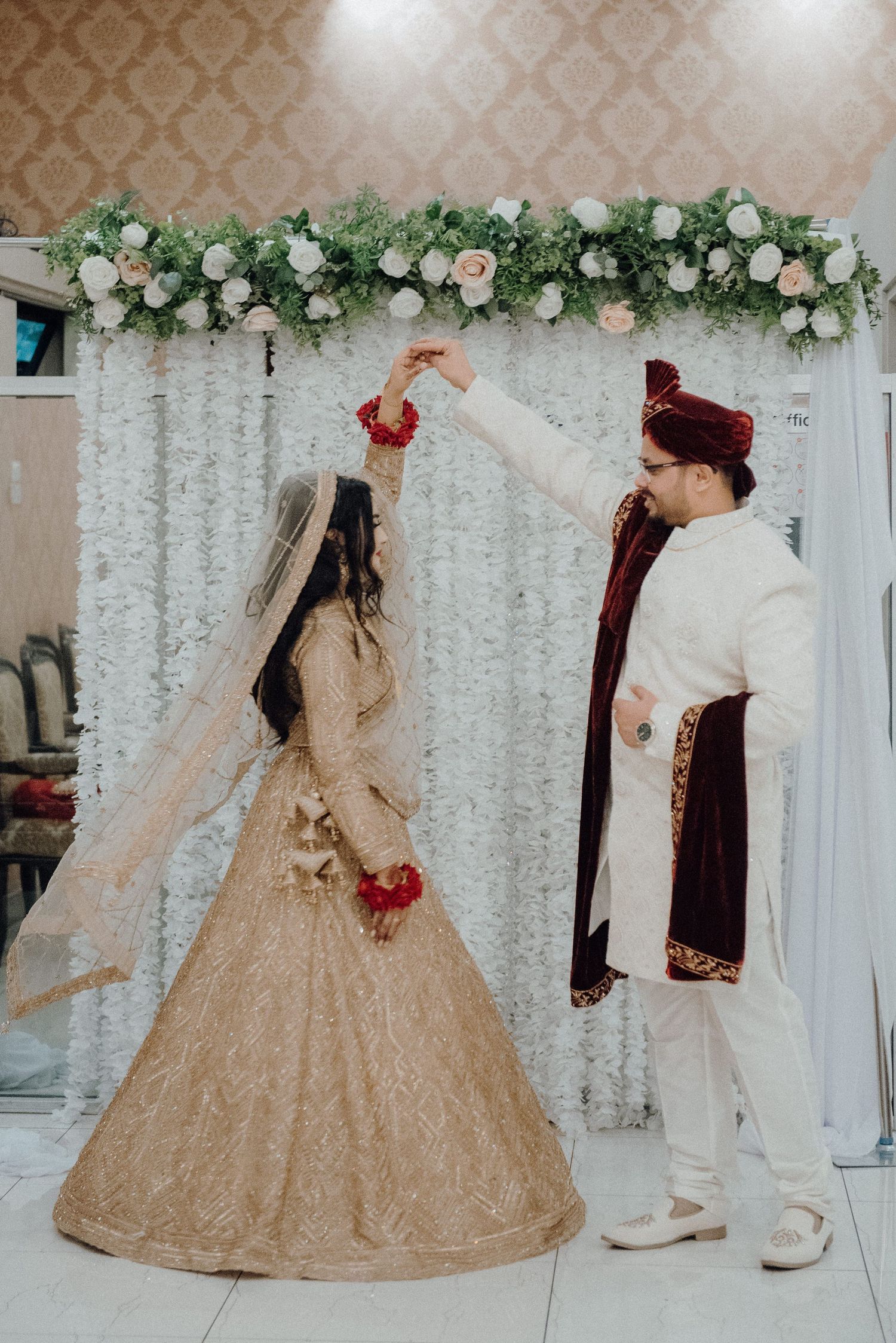Wedding couple in traditional South Asian attire dancing under floral arch.