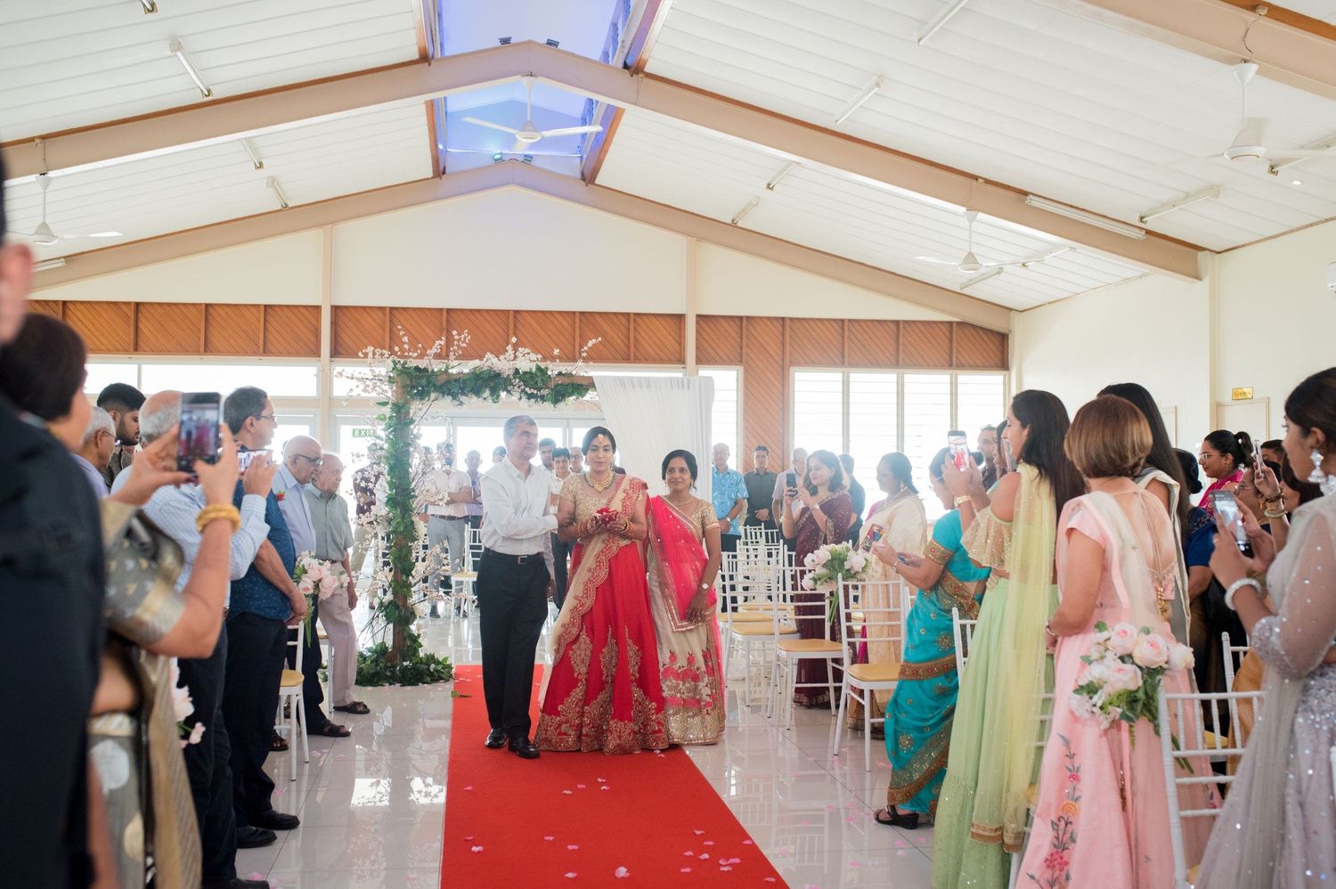 Wedding guests line a red carpet aisle in an elegant white venue as couple walks down during ceremony.