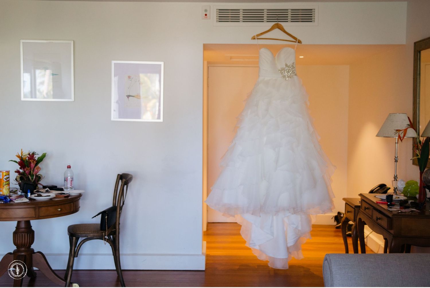 An elegant white wedding gown displayed on a hanger against a peach-colored wall.