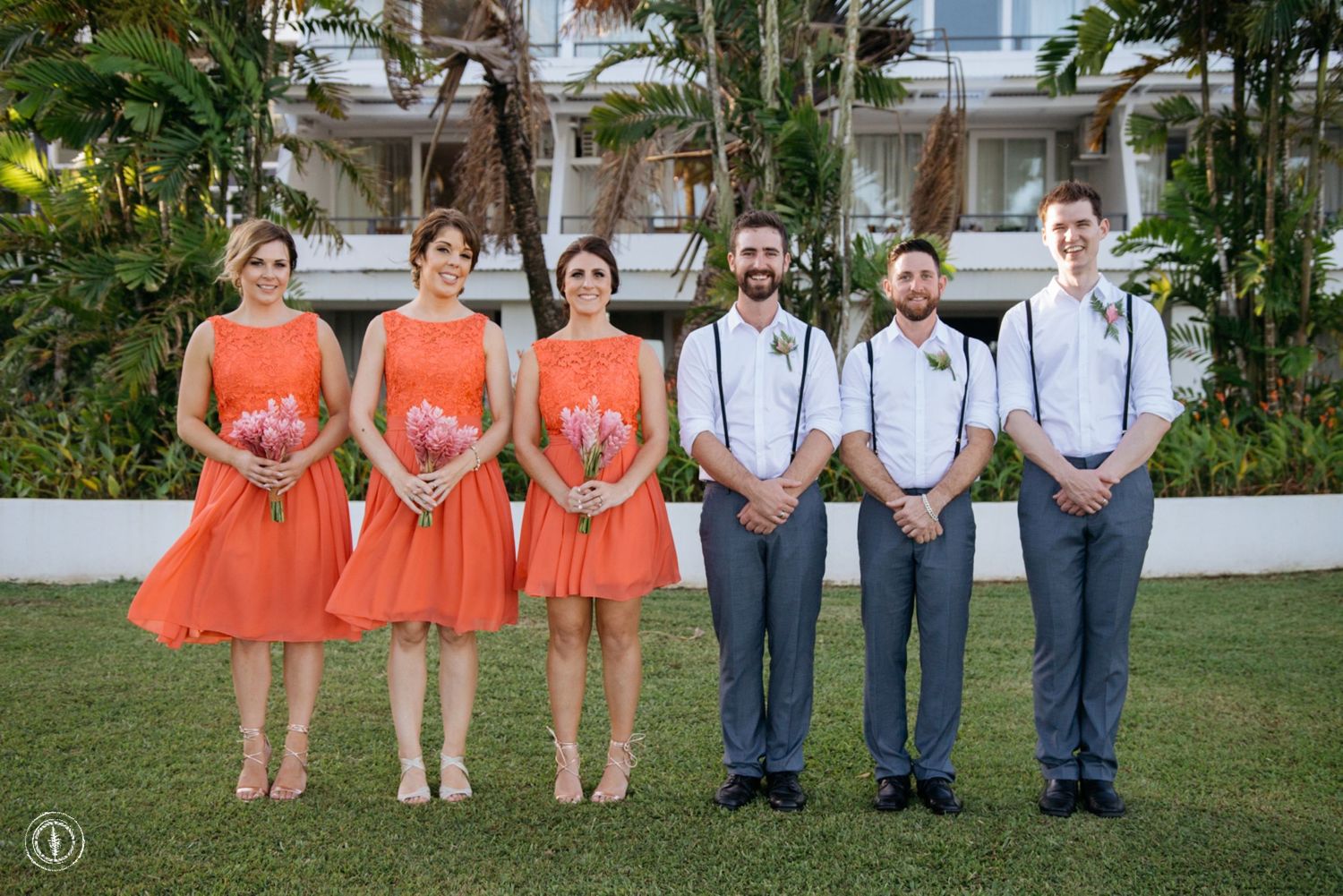 Wedding party in orange dresses and gray suits posing on a tropical lawn.