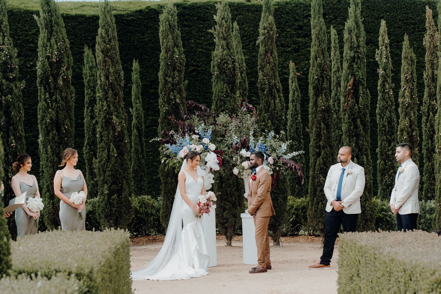 Wedding ceremony in progress between rows of cypress trees with attendants standing by.