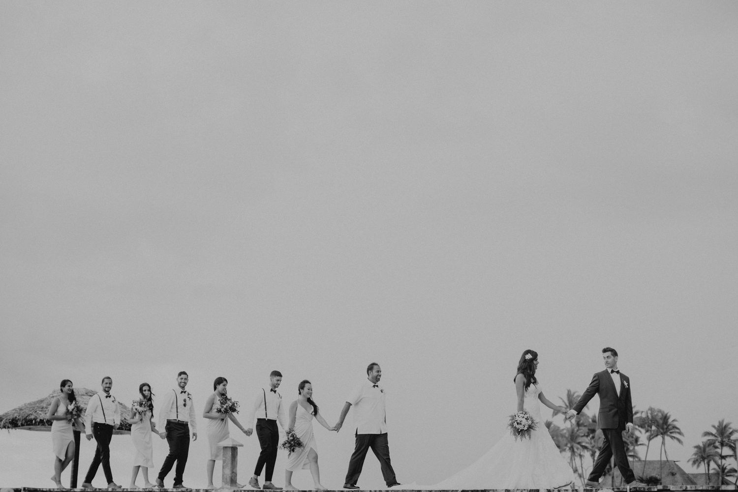 Wedding party walks in a line across a beach horizon in a stunning black and white photograph.