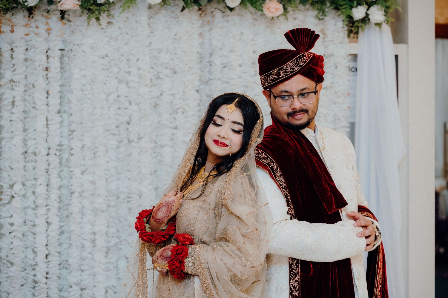 Newlyweds in coordinating traditional attire smile together against decorated backdrop.
