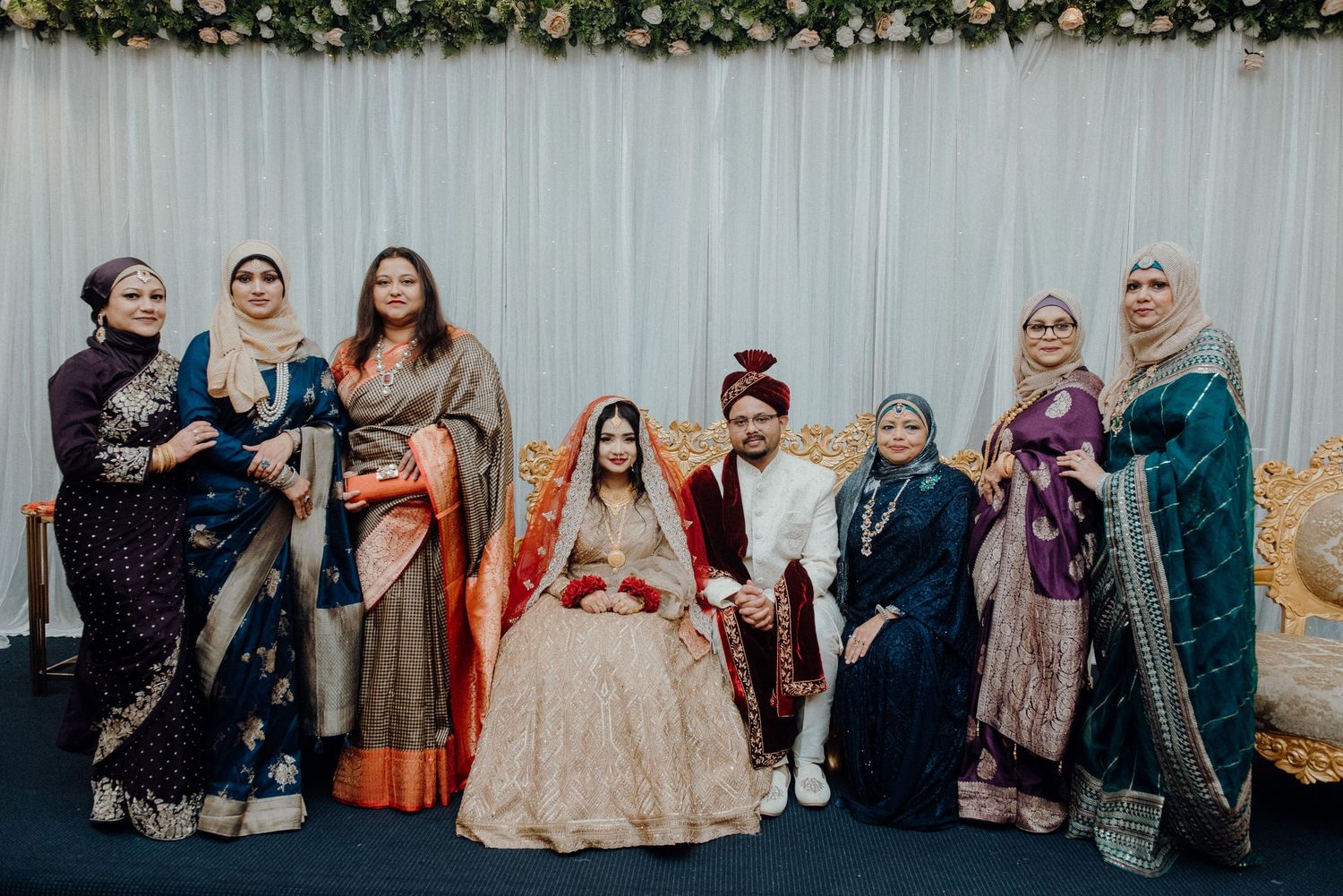 Traditional South Asian wedding group photo with ornate saris and formal attire against white backdrop.