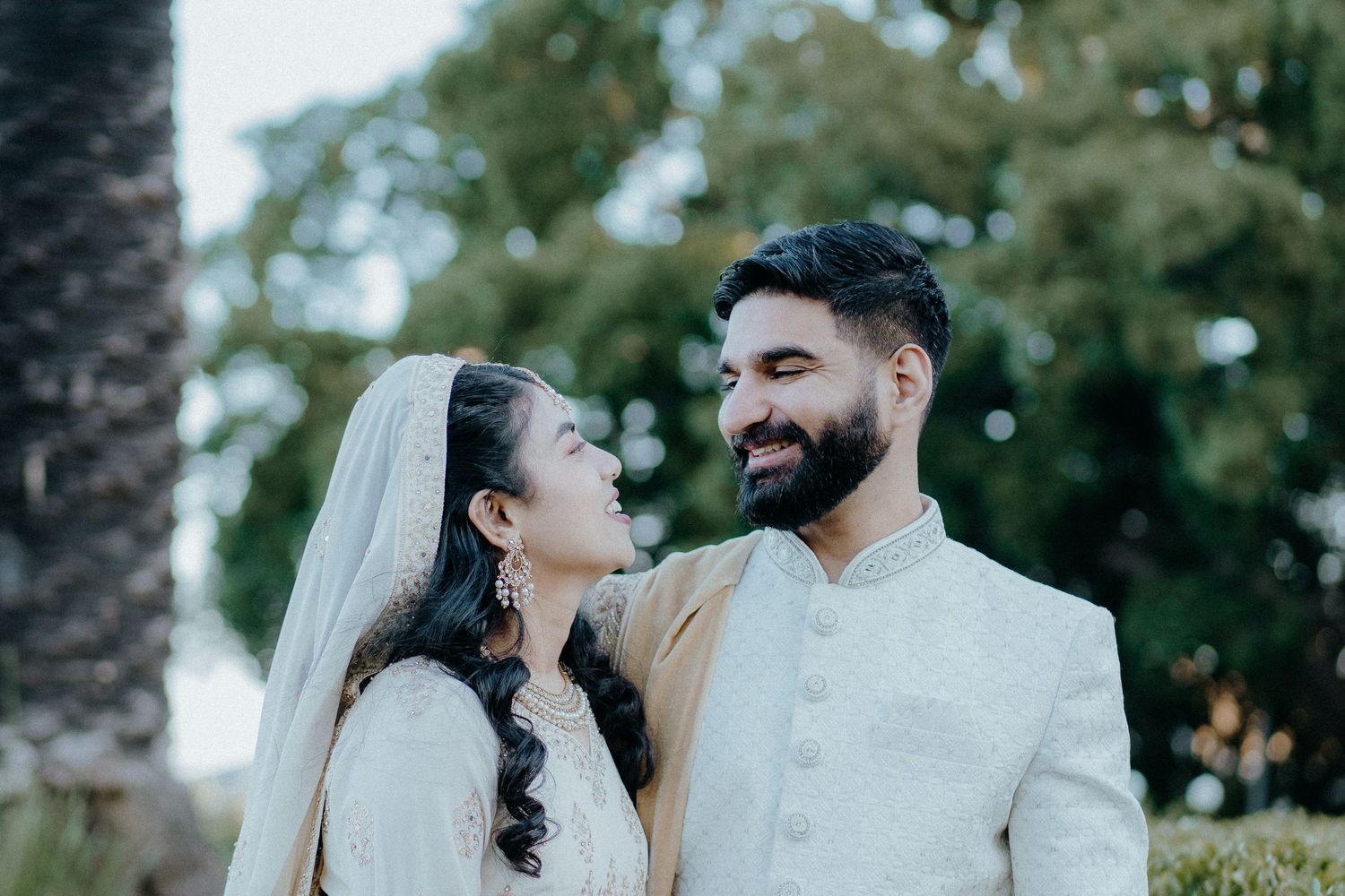 A couple in traditional cream and white wedding attire share a romantic moment outdoors.