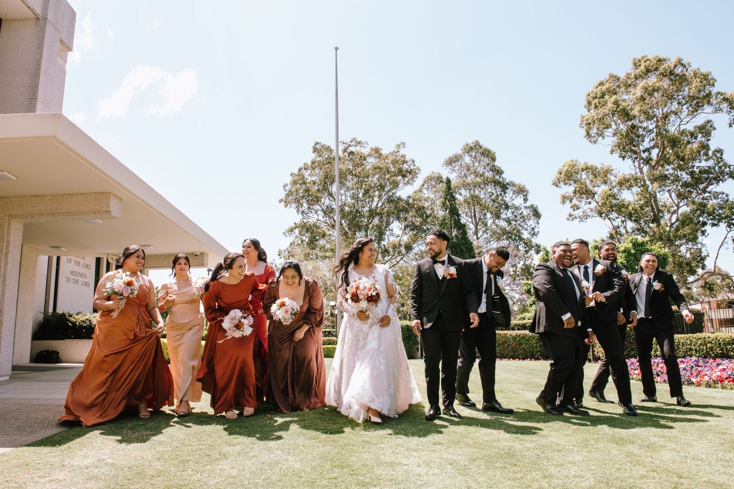 Wedding party walking outdoors on green lawn with bridesmaids in rust dresses and groomsmen in black suits.