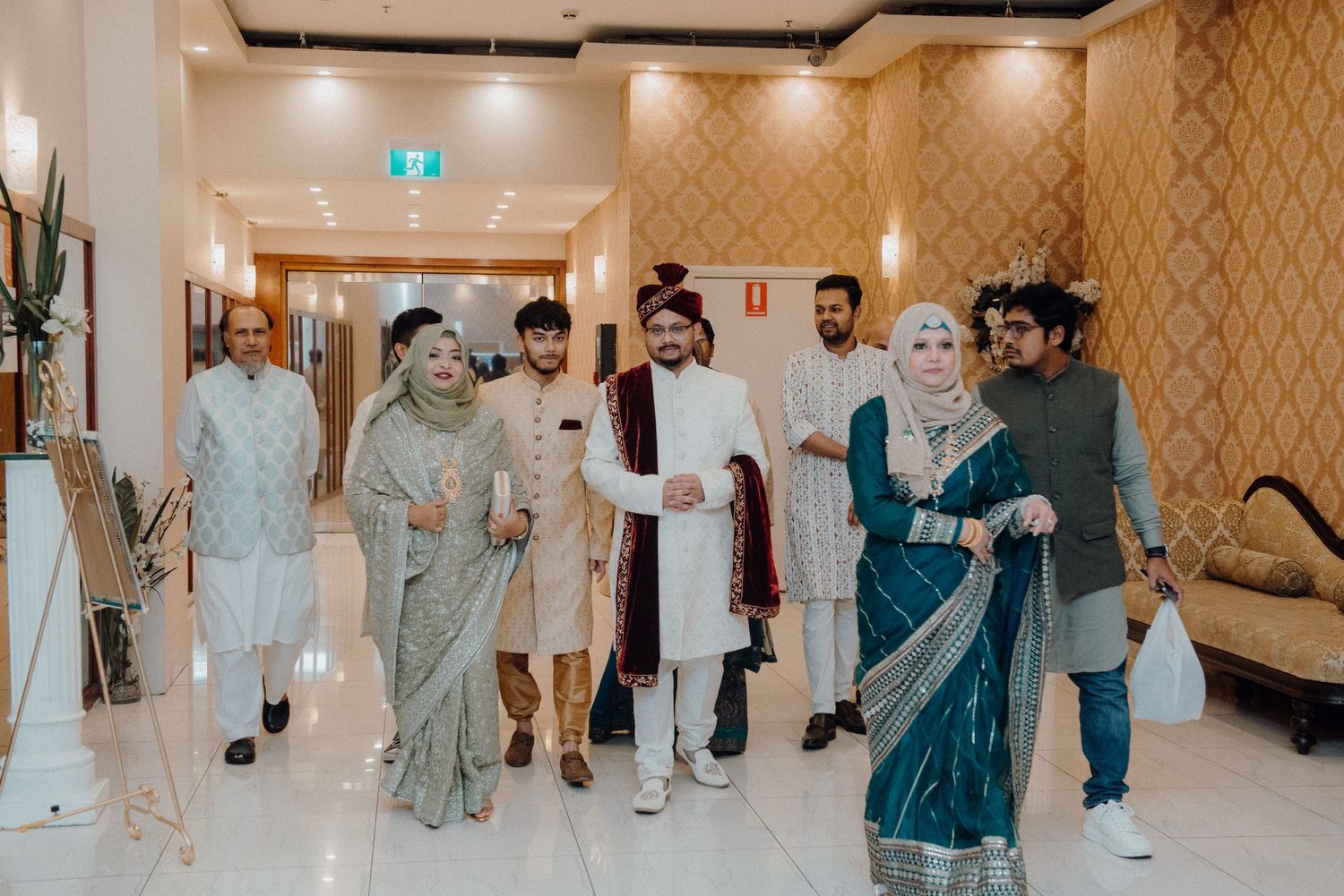 A group celebrates in traditional South Asian formal wear walking through an elegant hallway.