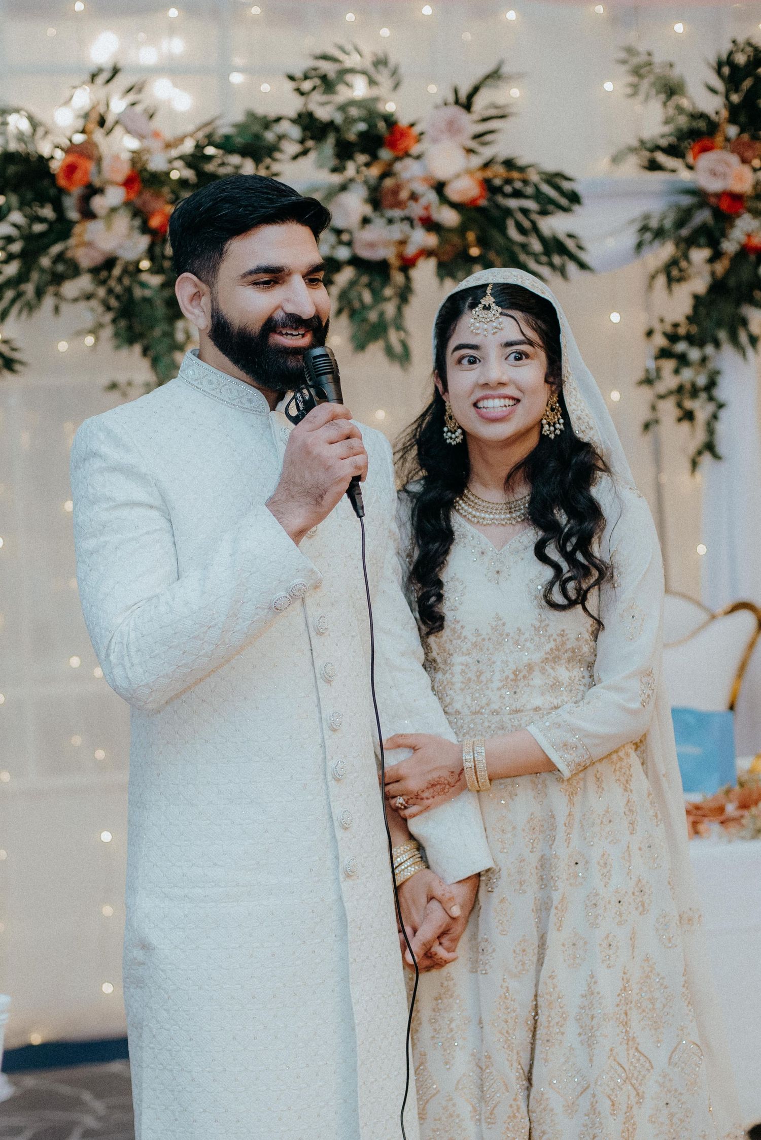 Wedding couple sharing a moment with microphones against a decorated backdrop with string lights.