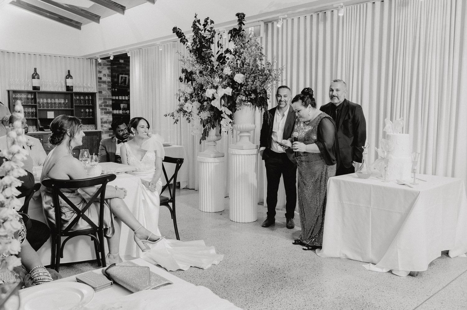 Black and white photo of wedding reception with elegant floral arrangements and guests.