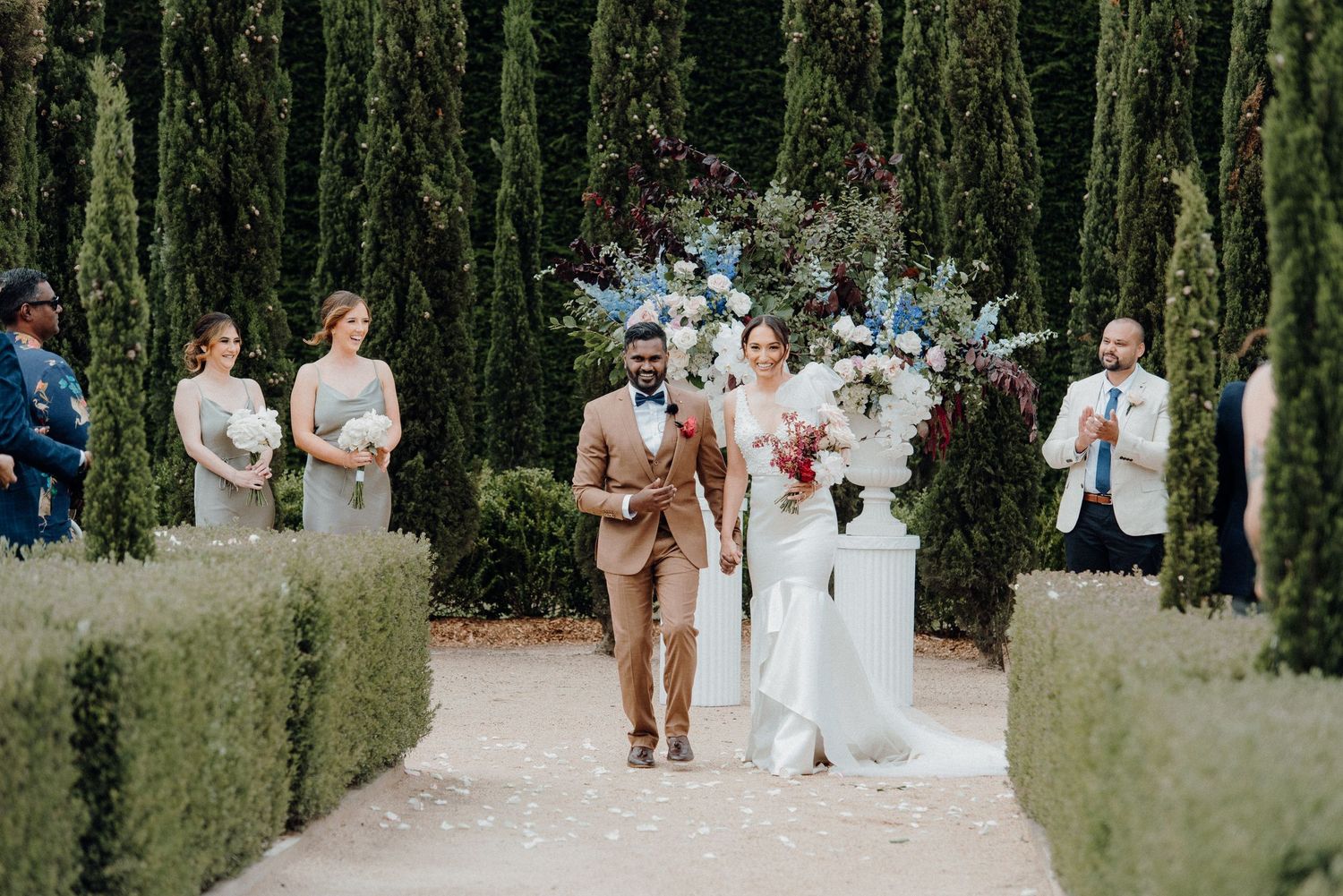 Wedding couple walking down path lined with tall cypress trees and manicured hedges.
