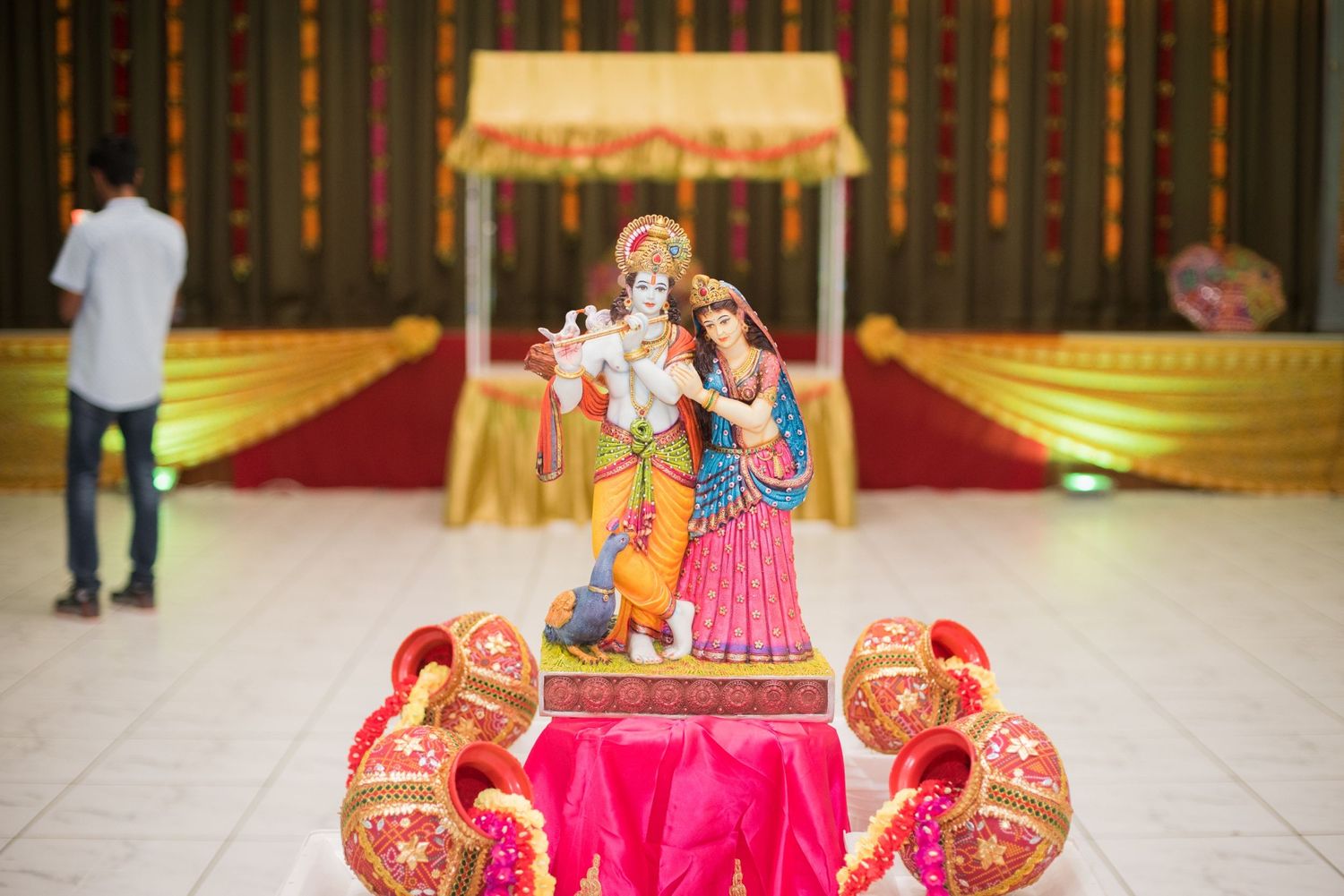 Decorative Krishna and Radha idol display with pink and gold decor at an Indian wedding ceremony.