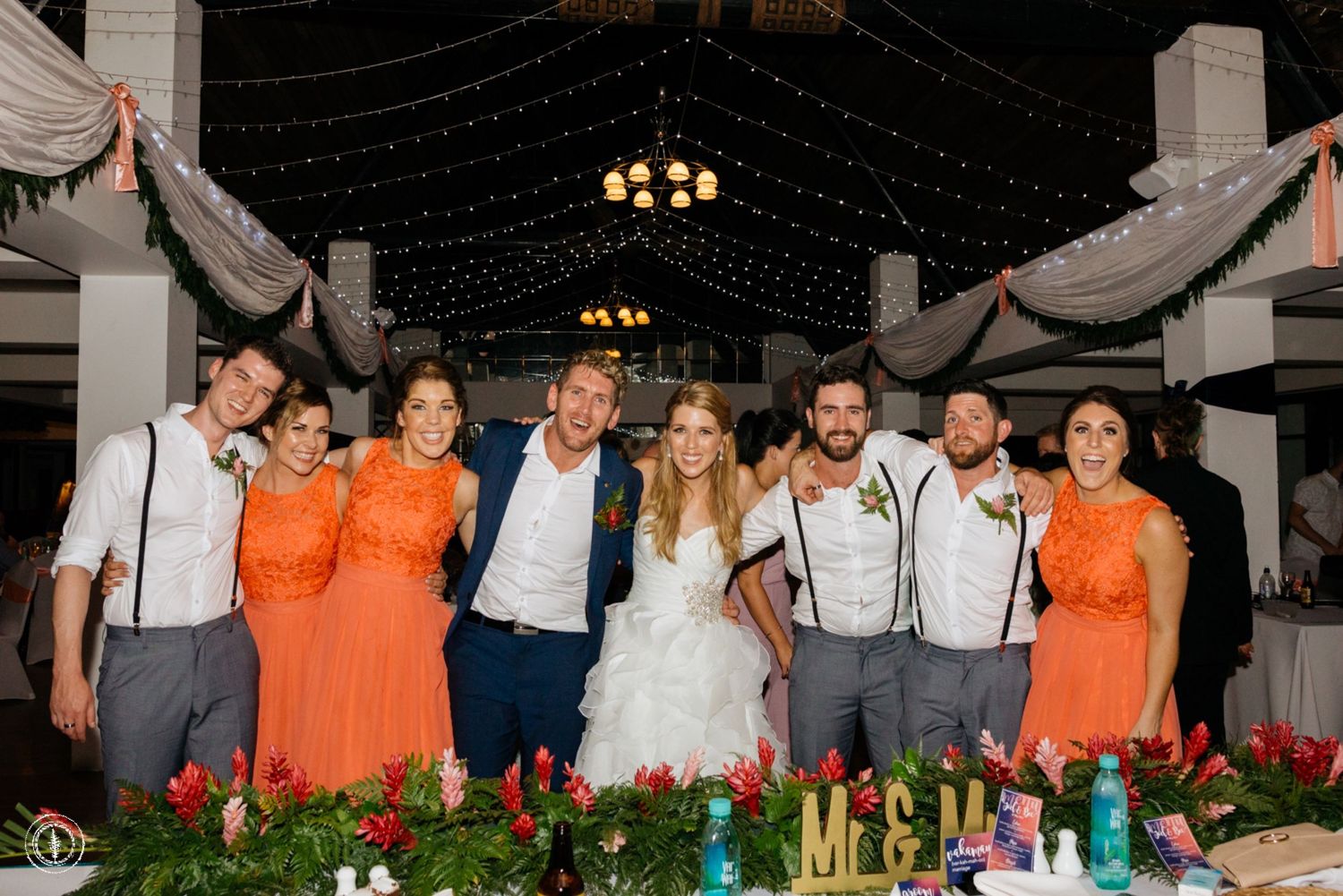 Wedding party in coral orange dresses and gray suspenders posing with greenery decorations.