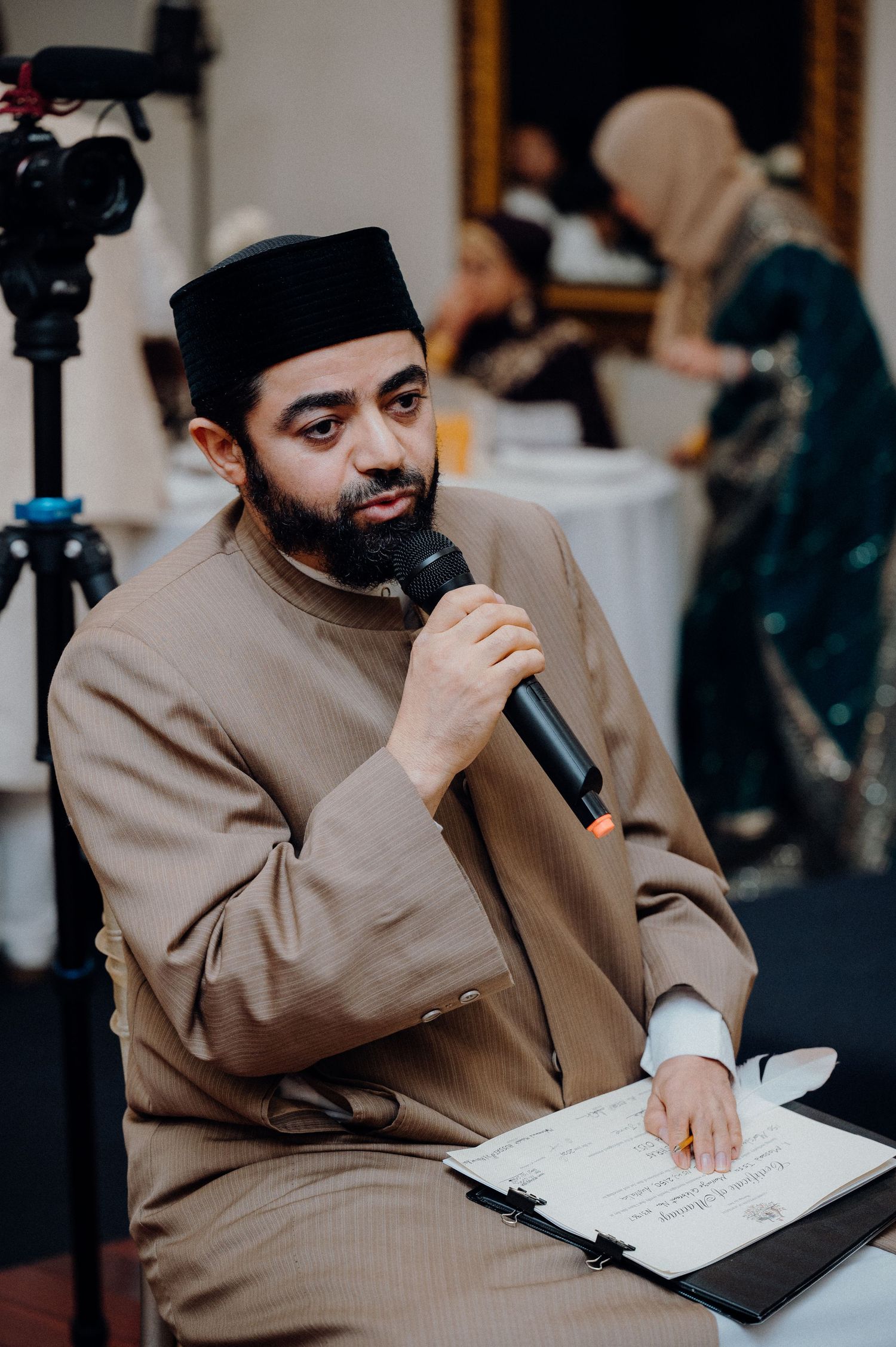 Speaker holding microphone at formal event wearing traditional cap and brown attire.