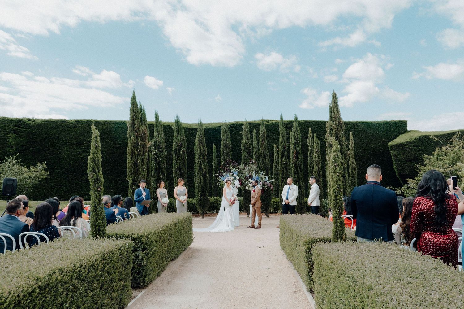 Outdoor wedding ceremony taking place between tall cypress trees with guests seated on both sides.