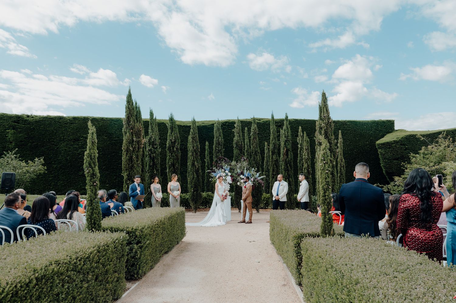 Elegant outdoor wedding ceremony in a formal garden with tall cypress hedges and guests seated along a manicured pathway.