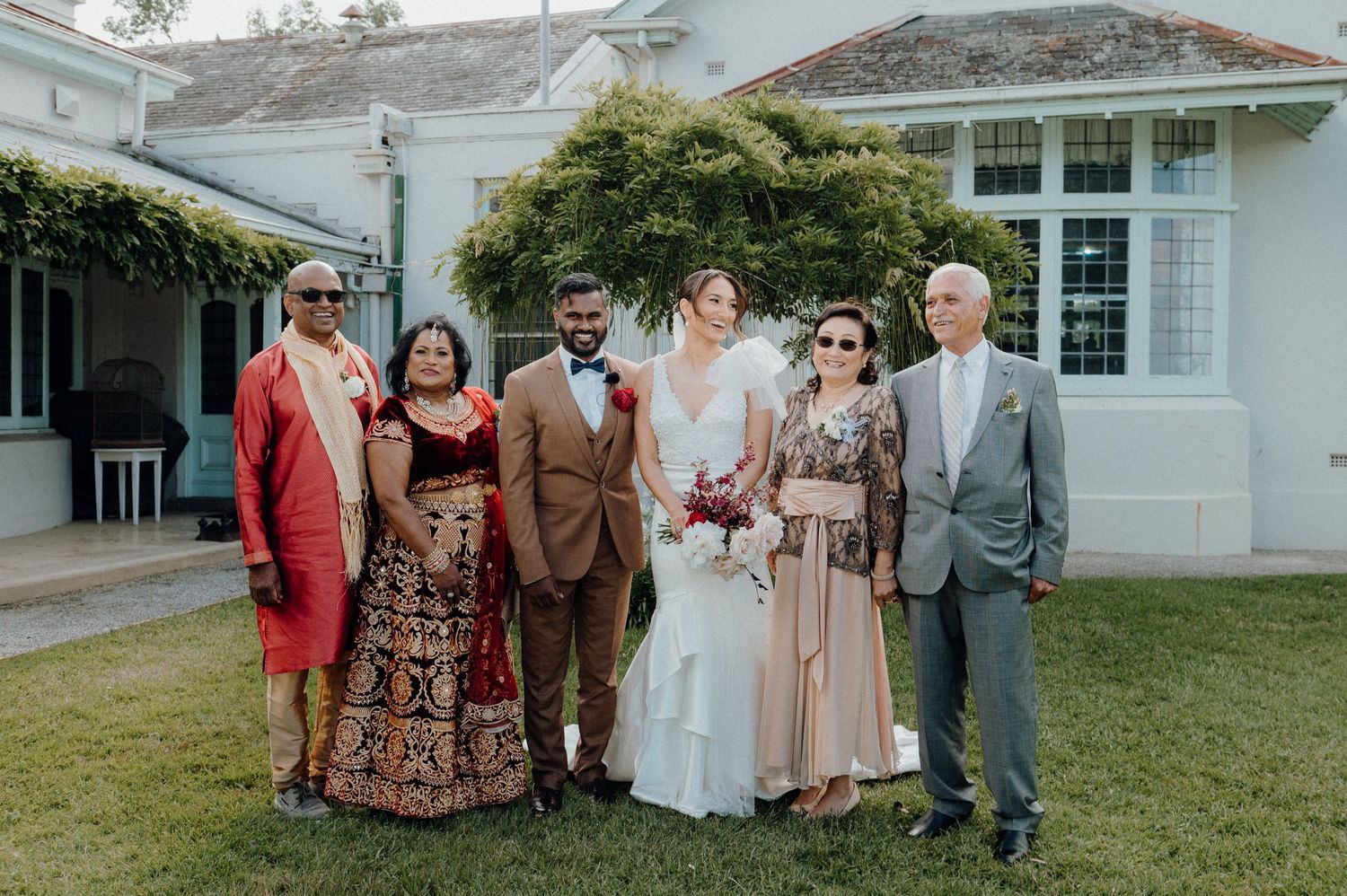 Wedding party posing for a formal photo outside a white colonial-style building with a garden backdrop.
