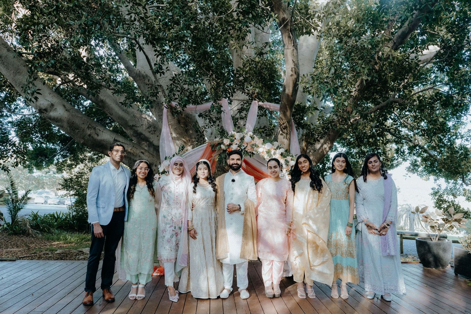 Wedding party group photo in pastel outfits under a large tree on wooden deck.