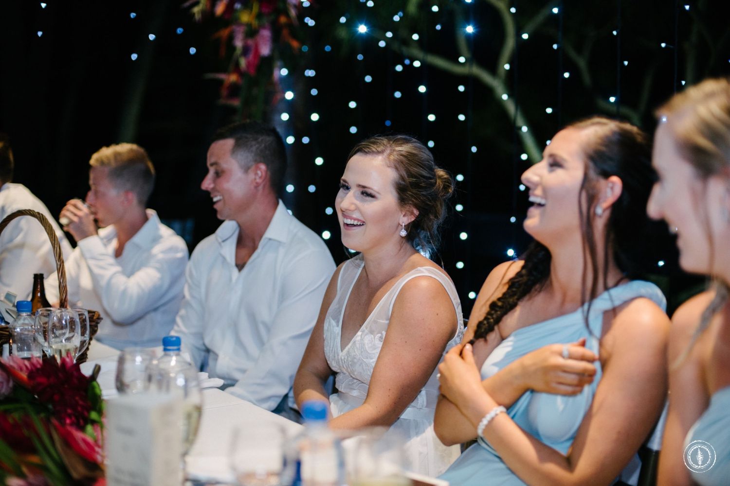Wedding party reacting with laughter during reception speeches under twinkle lights.