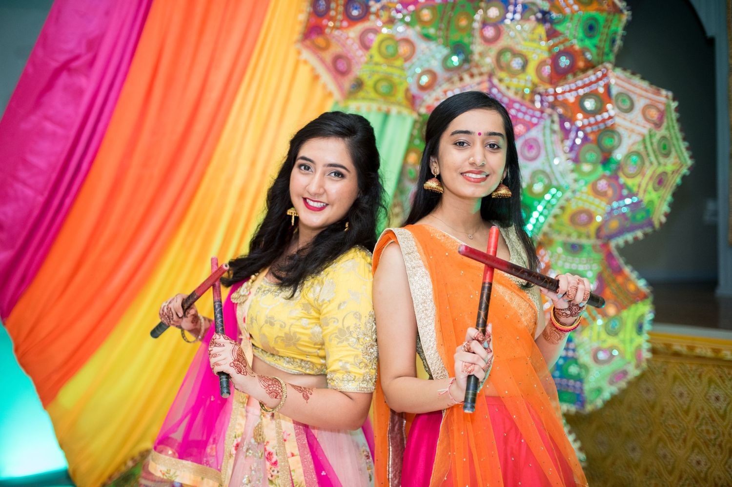 Two women in bright lehenga outfits posing with umbrellas against a colorful backdrop.