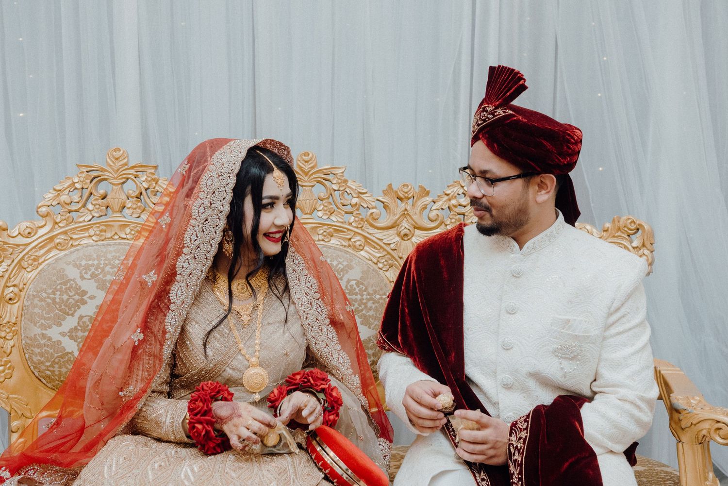 Couple in traditional South Asian wedding attire sharing a moment on decorative golden throne.