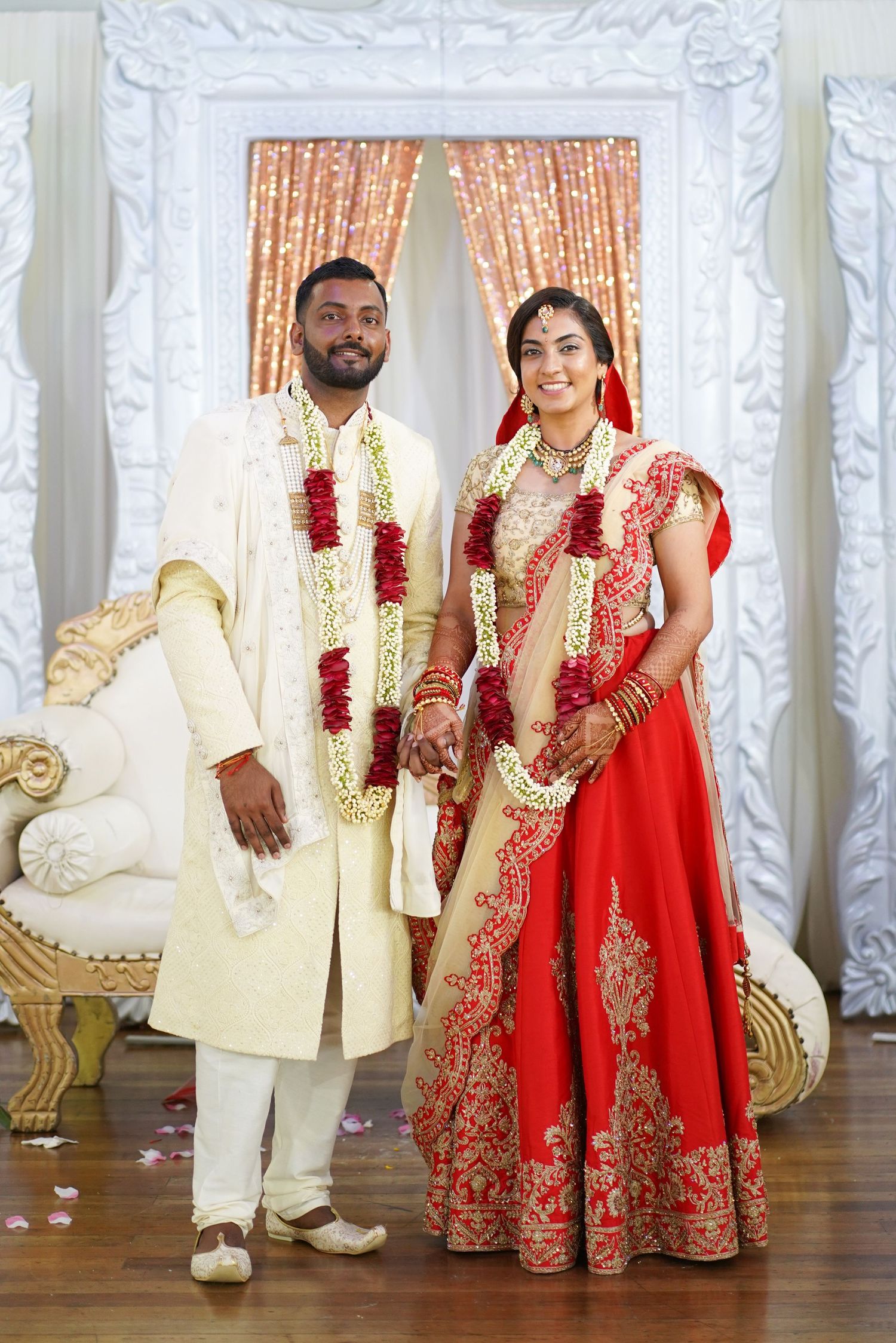 Couple in traditional Indian wedding attire standing against white backdrop with curtains.