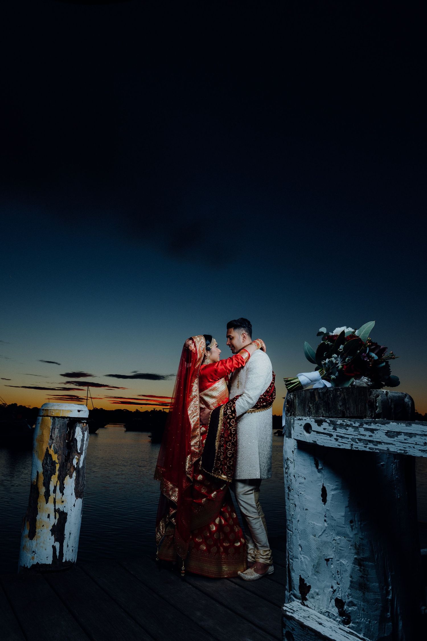 A couple in traditional wedding attire embracing on a dock at sunset.