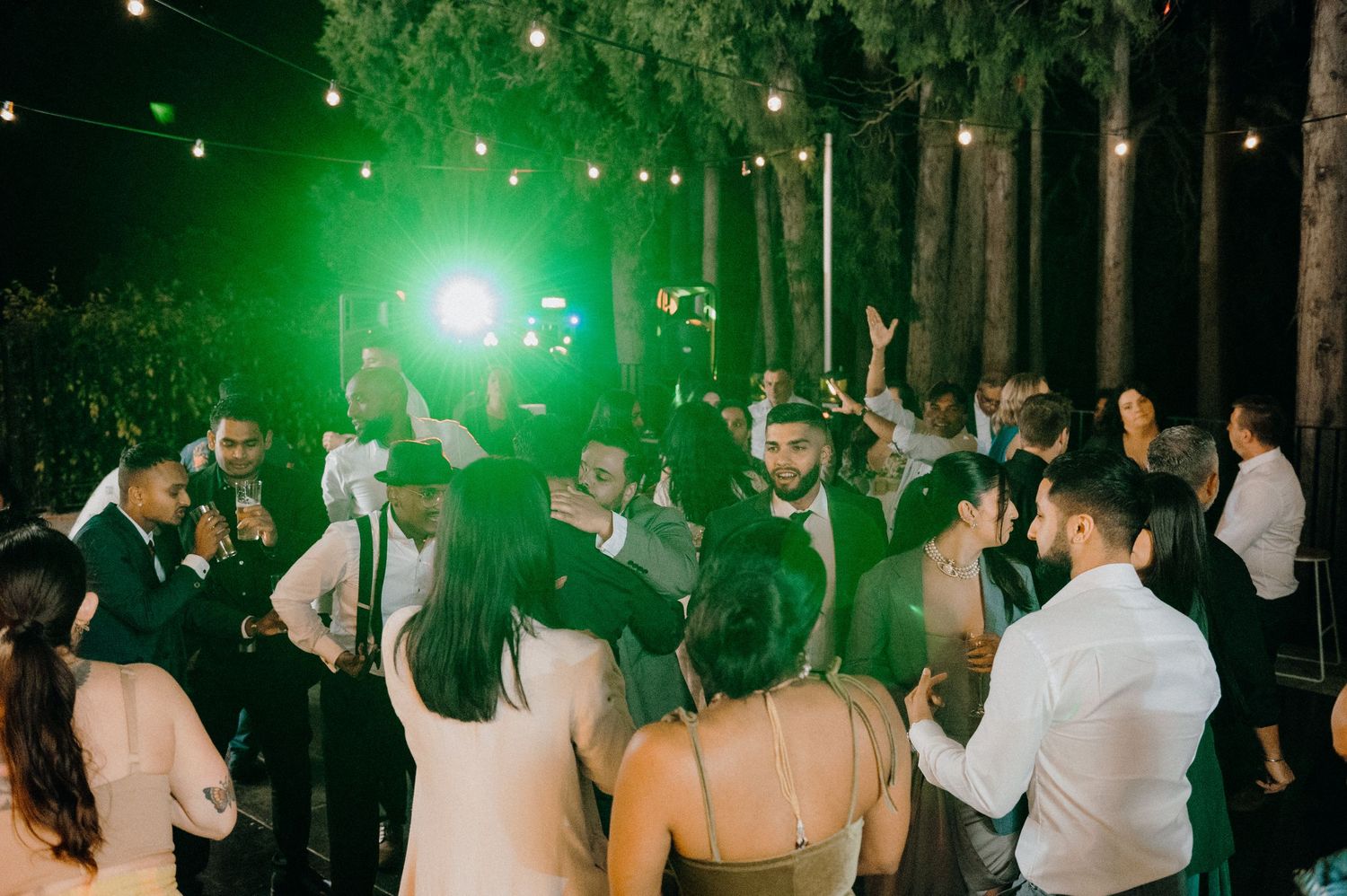 Wedding reception dance floor with green lighting and guests dancing under string lights.