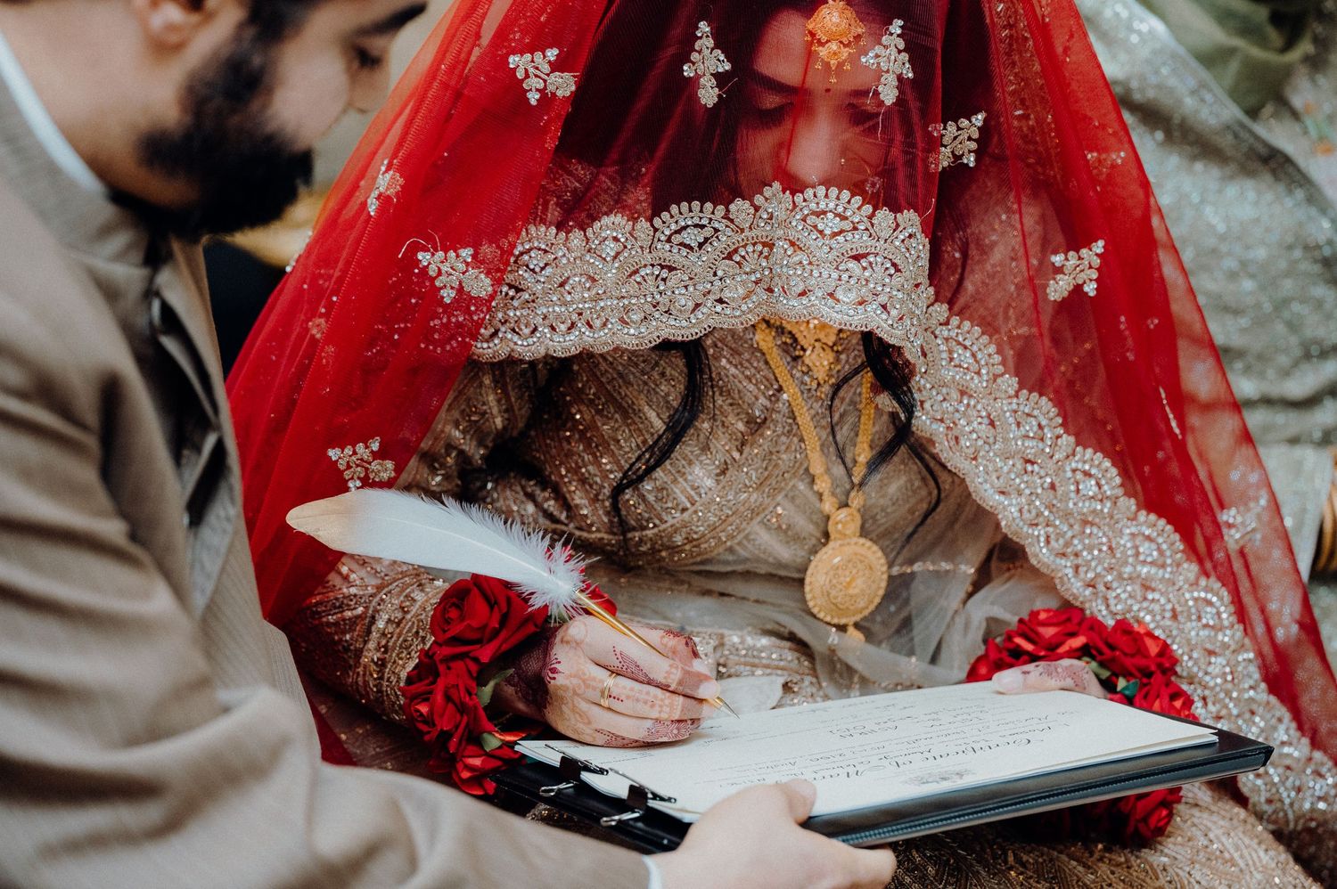 Wedding ceremony signing moment with traditional red bridal veil and document on clipboard.